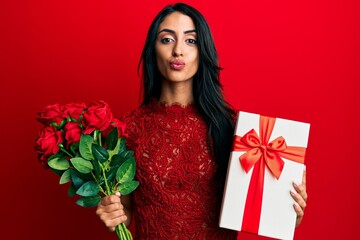 Beautiful hispanic woman holding anniversary gift and roses bouquet looking at the camera blowing a kiss being lovely and sexy. love expression.