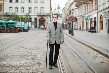 Senior man in stylish outfit walking on street with cane in hand. Caucasian pensioner with grey...