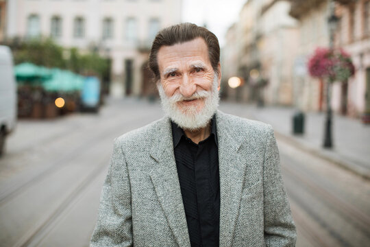 Portrait Of Happy Man With Grey Beard Mature Male In Grey Jacket And Black Shirt Smiling And Looking At Camera. Blur Background Of Big City Street.