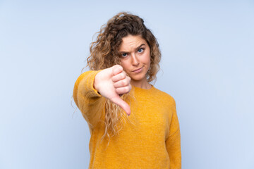 Young blonde woman with curly hair isolated on blue background showing thumb down with negative...