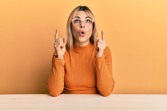 Young caucasian woman wearing casual clothes and glasses sitting on the table amazed and surprised looking up and pointing with fingers and raised arms.