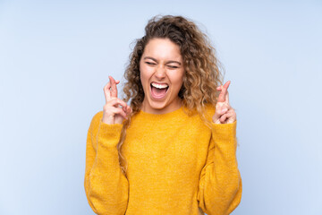 Young blonde woman with curly hair isolated on blue background with fingers crossing