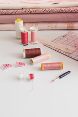 Three laid focused thread reels surrounded by more tailor accessories on a white table with a pile of folded pinkish fabrics as background.