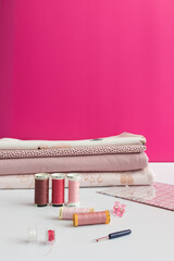 Folded pinkish fabrics piled next to several thread reels and more tailor accessories on a white table with a pink background.