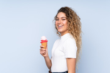 Young blonde woman with curly hair holding a cornet ice cream isolated on blue background smiling a lot