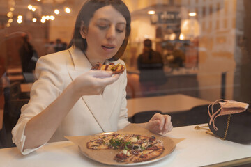 Young adult woman having pizza bite at the restaurant. Photo through the window glass. Warm toned colored filter