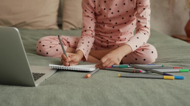 Cute little African American girl learning to draw with felttip pens by looking to laptop. Teenage girl in pajamas sits on bed in bedroom with beautiful light interior. Slow motion. Close up.