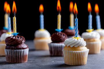 Close up side view image of an assortment of chocolate and vanilla cupcakes with white and brown frostings on. Each have a candle lit against dark background. Celebration, party, birthday concepts.