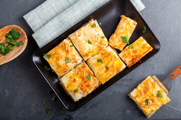 Turkish kiymali borek served on a tray after being sliced into square shapes. Fresh parsley leaves are sprinkled on top for added flavor. One slice is on a spatula for serving. Made with phyllo dough
