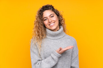 Young blonde woman with curly hair wearing a turtleneck sweater isolated on yellow background presenting an idea while looking smiling towards