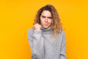 Obraz na płótnie Canvas Young blonde woman with curly hair wearing a turtleneck sweater isolated on yellow background with angry gesture
