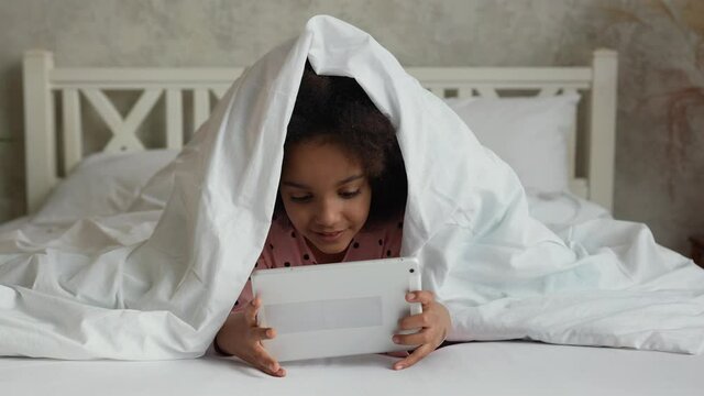 Portrait little African American girl watches cartoon on digital tablet, gets scared and hides under blanket with his head. Teenage girl in pajamas lies on white bed in bedroom. Slow motion. Close up.