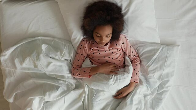 Top view of a little sleeping African American girl waking up stretching and yawning. Teenage girl in pajamas lies on white bed in bedroom. Slow motion.