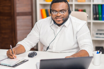 Fototapeta premium Workplace of freelancer. African-American man works at home office using computer, headset and other devices. Employee is having a conference call. Remote job.