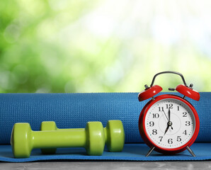 Alarm clock, yoga mat and dumbbells on grey marble table outdoors. Morning exercise