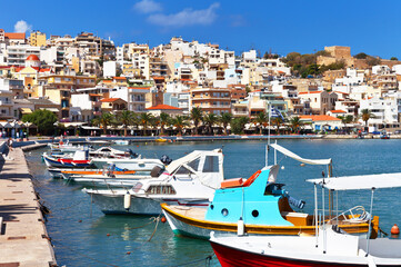 Greece island Crete. Beautiful view of the town of Sitia on the hill and fishing boats on blue water near embankment on sunny day. Southern bright seascape. Summer seaside vacation and travel