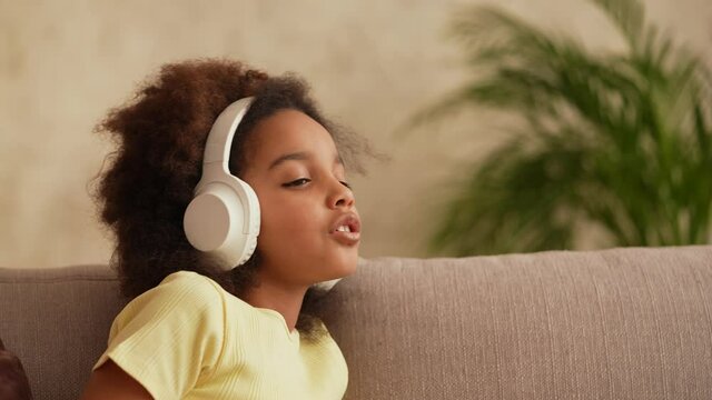 Portrait little African American girl in big white wireless headphones listens to music. Teenage girl sits on gray sofa in hall against backdrop of bright beautiful room interior. Slow motion.
