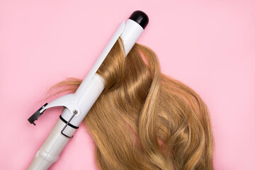Curling blonde hair on a large diameter curling iron on a pink background. Hair health concept, damage by hot hair styling.