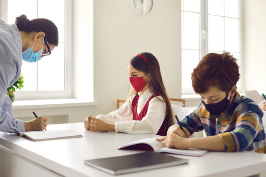 Lesson at new normal classroom, back to school after coronavirus quarantine lockdown. Teacher in face mask writing notes in pupil notebook while boy classmate performing task sitting next to girl