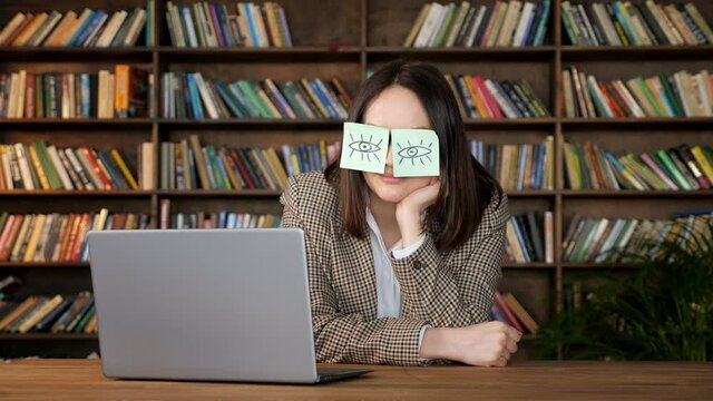Brunette with short hair takes nap, holding head on hand with eye painting stickers on face near laptop on brown wooden table in office with books