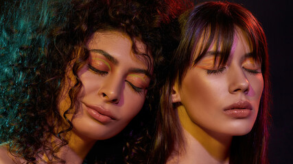 Beauty portrait of two young brunette female models with professional art makeup posing with eyes closed in neon light isolated over black background