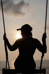 Photo silhouette of a woman wearing a hat sitting on a swing with the evening sunshine.
