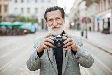 Handsome man with grey beard holding old fashioned photo camera while standing outdoors. Blur background of old city street. Travelling concept.