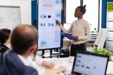 Black woman worker explaning company statistics in front of colleagues, briefing group of...