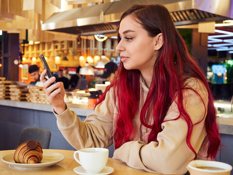 Young beautiful woman with long red hair uses a mobile phone in a restaurant interior with a cup of coffee and a croissant.
