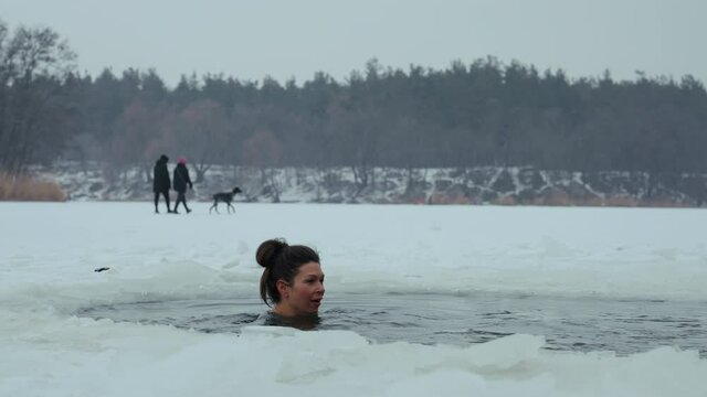 Woman with hair bun plunging in ice hole in frozen river, blurred people walking with dog on background. Young female enjoying winter traditions. Concept of extreme activities