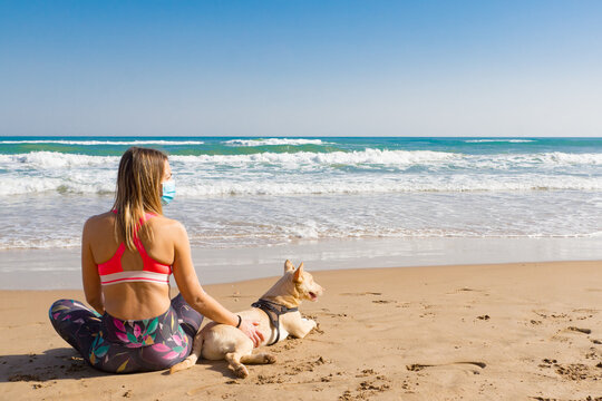 A Fitness Girl Is Sitting On The Seashore With A Dog And Wearing A Covid Mask