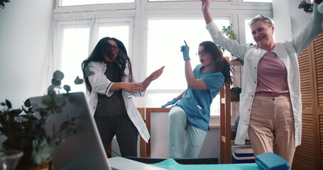 Three fun happy multiethnic female doctors dance together at work lab celebrating victory over coronavirus pandemic.