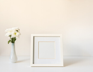 Closeup of blank picture frame next to white chrysanthemums in small vase on table against wall...