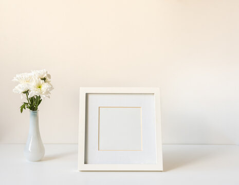 Closeup of blank picture frame next to white chrysanthemums in small vase on table against wall (selective focus)