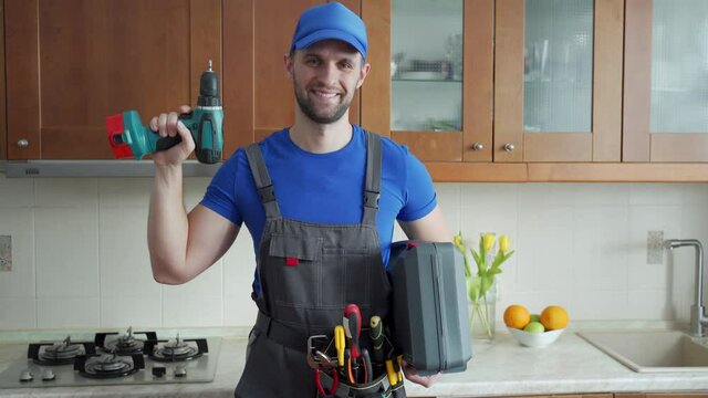 Repairman holds a screwdriver and a suitcase of tools in the kitchen and looks at the camera