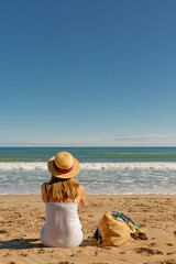A girl with a hat is sitting on the sand and looking to the sea with her bag 