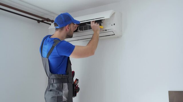 Male technician in overalls and a blue cap repairs an air conditioner on the wall