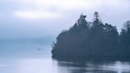 Dramatic landscape image looking across Derwentwater in Lake District towards Catbells snowcapped mountain with thick fog rolling through valley