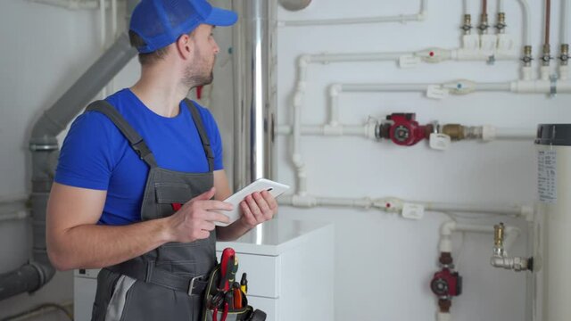 Young worker in a protective suit uses a tablet while checking the boiler equipment in the house.