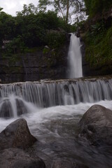 A stunning jungle waterfall flows into a cool mountain river with big rocks.