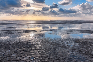 Sunset on the Wadden Sea in The Netherlands during low tide and cracked ground in the foreground. Dramatic seascape with clouds and reflections in blue and orange tones.