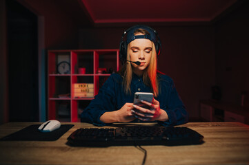 Beautiful girl gamer uses a smartphone while playing on the computer with a serious face. Female gamer in a headset sits at a table at the computer and uses the Internet on a smartphone.