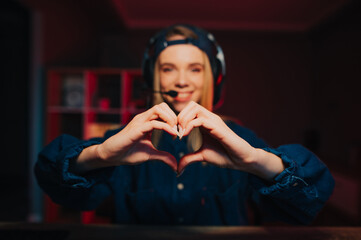 Female streamer in headset shows heart gesture and smiles at camera. Background. Gamer woman's hands show heart.