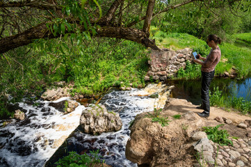Naklejka na ściany i meble A mature woman on the bank of a small river photographs the landscape with a tablet. The remains of a hydraulic structure form a waterfall.