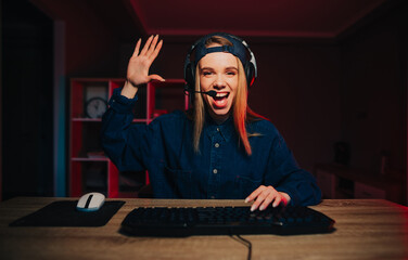 Positive female streamer sits at night in the room at the computer and communicates on a headset with subscribers, poses palms of greetings to the camera and smiles. © bodnarphoto