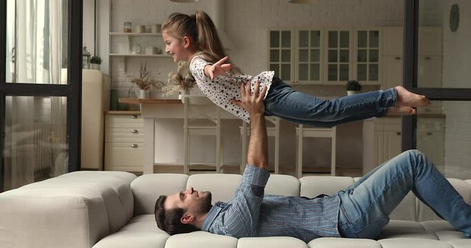 Strong dad lying on comfy couch in living room lifts up on outstretched arms his pretty little daughter, girl spread hands like wings of plane imagines fly in air having fun with loving father at home