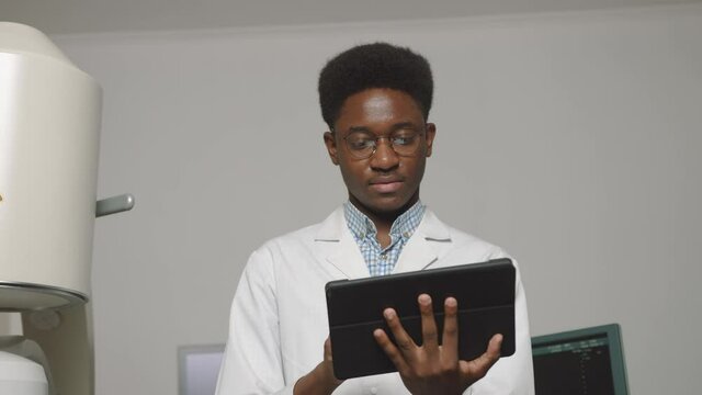 Close up portrait of smiling handsome African Doctor holding digital tablet pc.doctor, standing in front of ultrasound and lithotripter machines. Safety non-invasive stone treatment.
