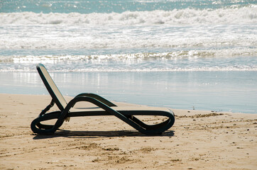 Plastic beach lounger stands on sandy beach at water's edge against backdrop of sea waves. Beach holiday concept, entrance by the sea. Space for text.