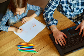 Top view of Father working in his home office on a laptop, her daughter sits next to her and draw