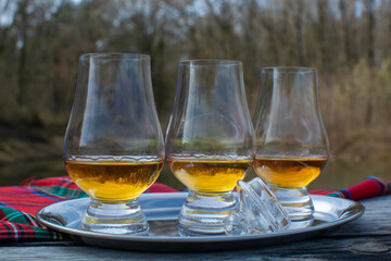 Tasting glasses of scotch whisky strong drink and red Scottisch tartan on old outdoor table with forest lake on background